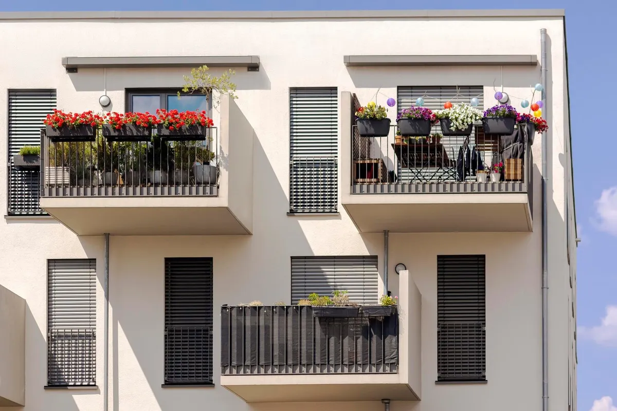 Façade d'immeuble récent à l'enduit clair, balcons fleuris et fenêtres fermées par des volets à lames gris foncé baissés