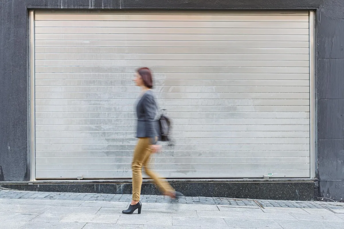 Rideau métallique à lames argentées fermé sur une devanture, devant lequel une passante floue marche sur le trottoir