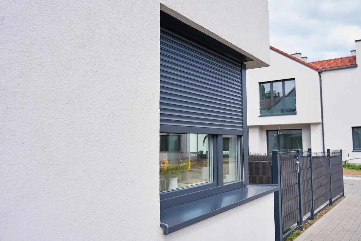 Fenêtre d'une maison récente à l'enduit blanc, volet roulant gris anthracite à moitié baissé, clôture grise et maisons voisines en arrière-plan